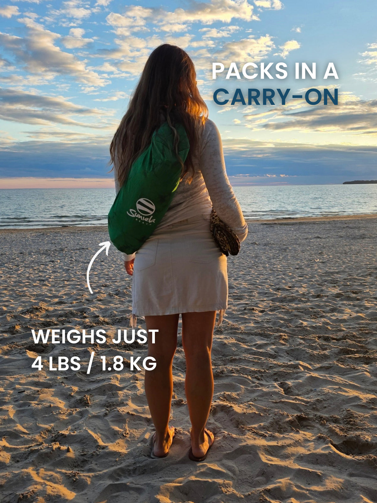 Woman standing on a beach at sunset carrying a Rainforest Green Suniela beach cabana in its compact shoulder bag. Packs in a carry-on and weighs just 4 lbs / 1.8 kg
