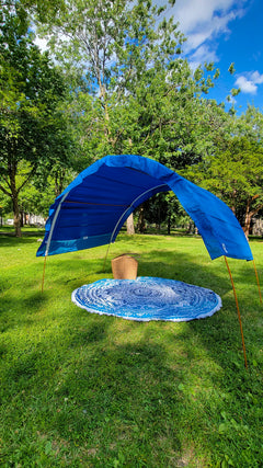 A blue canopy tent with a matching round blue mat set up on a green grassy lawn in a park with trees and a blue sky in the background.