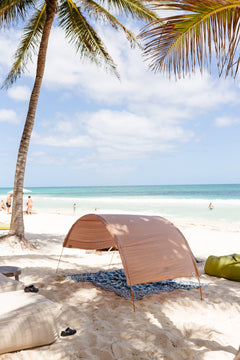 A tan beach canopy tent set up on white sand at a tropical beach, with a palm tree, ocean, and sky in the background.