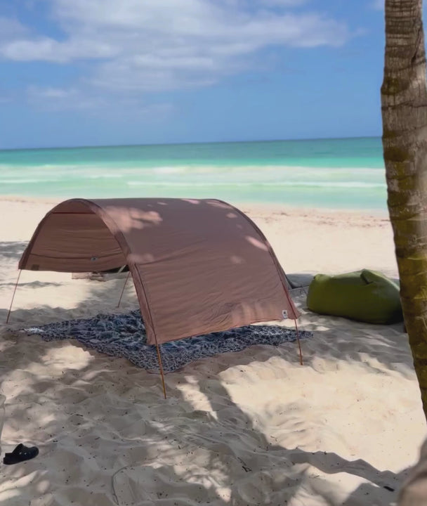 a video showing a sand coloured shade cabana by Suniela with a blue madala beach mat, on a sandy beach with turquoise water and blue sky in the background
