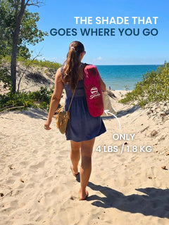 Woman carrying a pink Suniela portable beach cabana in its compact carry bag along a beach path, lightweight shade that goes wherever you go