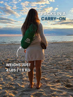 Woman standing on a beach at sunset carrying a Rainforest Green Suniela beach cabana in its compact shoulder bag. Packs in a carry-on and weighs just 4 lbs / 1.8 kg