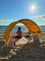 Man relaxing under a Tiger’s Eye Suniela portable beach cabana on a sunny beach, with an open view of the ocean through the hand-dyed cotton arch